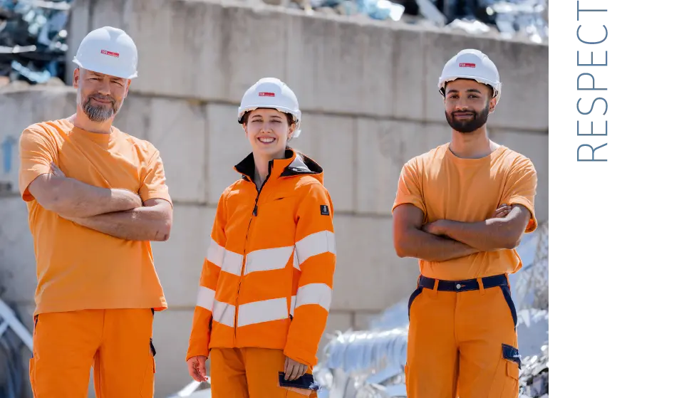  Three colleagues stand in protective gear in the scrapyard and look friendly into the camera
