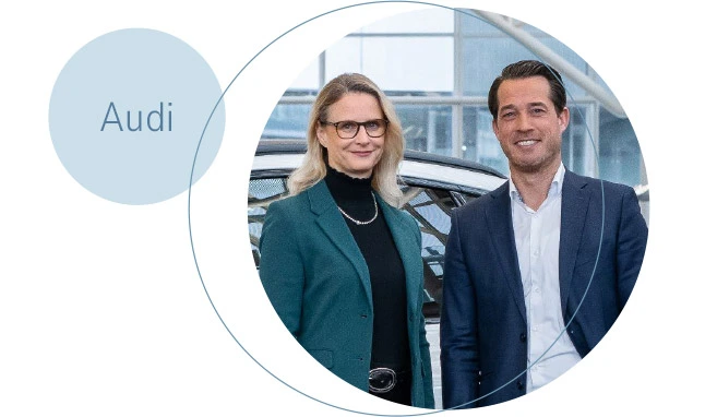 Eine Geschäftsführerin und ein Geschäftsführer vor einem Auto. Daneben ein Krei smit der Inschrift "Audi"