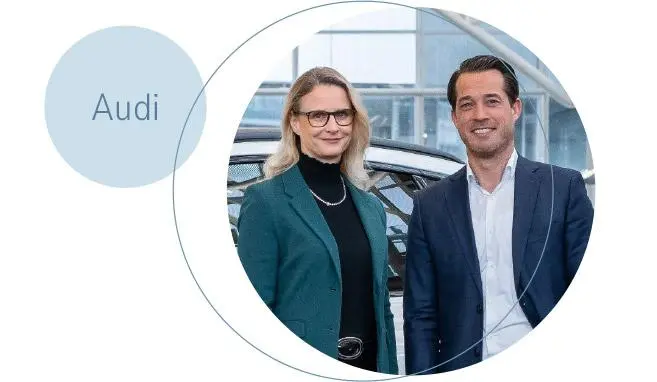 Eine Geschäftsführerin und ein Geschäftsführer vor einem Auto. Daneben ein Krei smit der Inschrift "Audi"