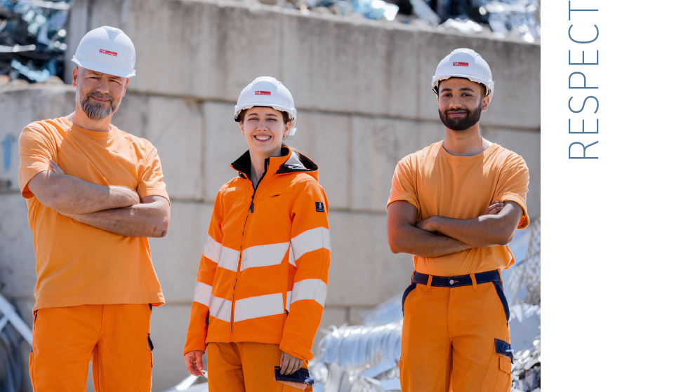  Three colleagues stand in protective gear in the scrapyard and look friendly into the camera